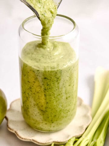 Celery smoothie being poured into a glass surrounded by ingredients.