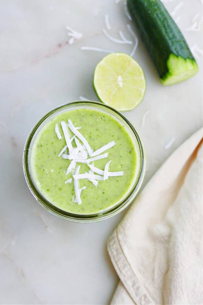 cucumber smoothie topped with shredded coconut flakes on a counter next to ingredients