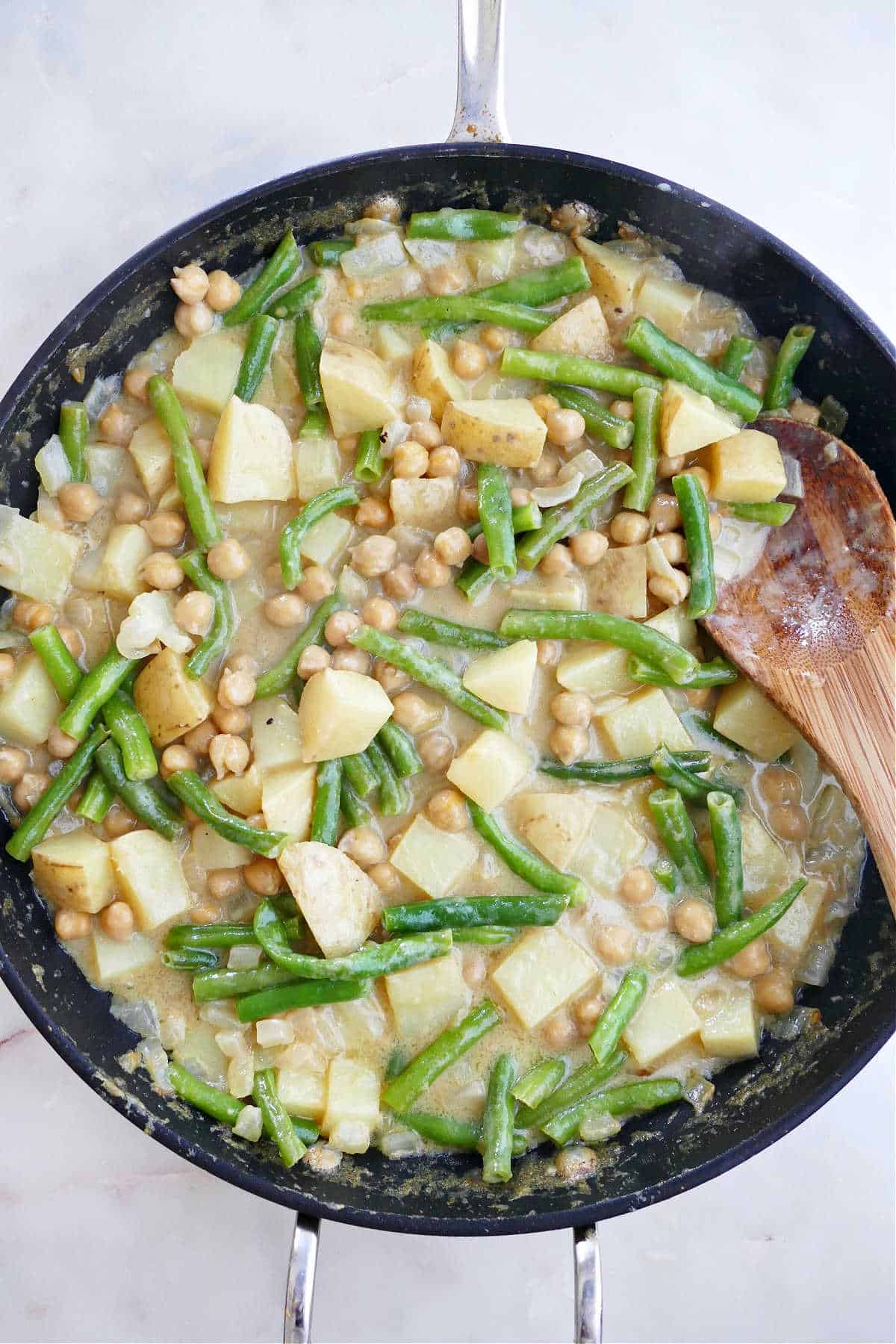 green bean curry cooking in a large skillet with a wooden spoon
