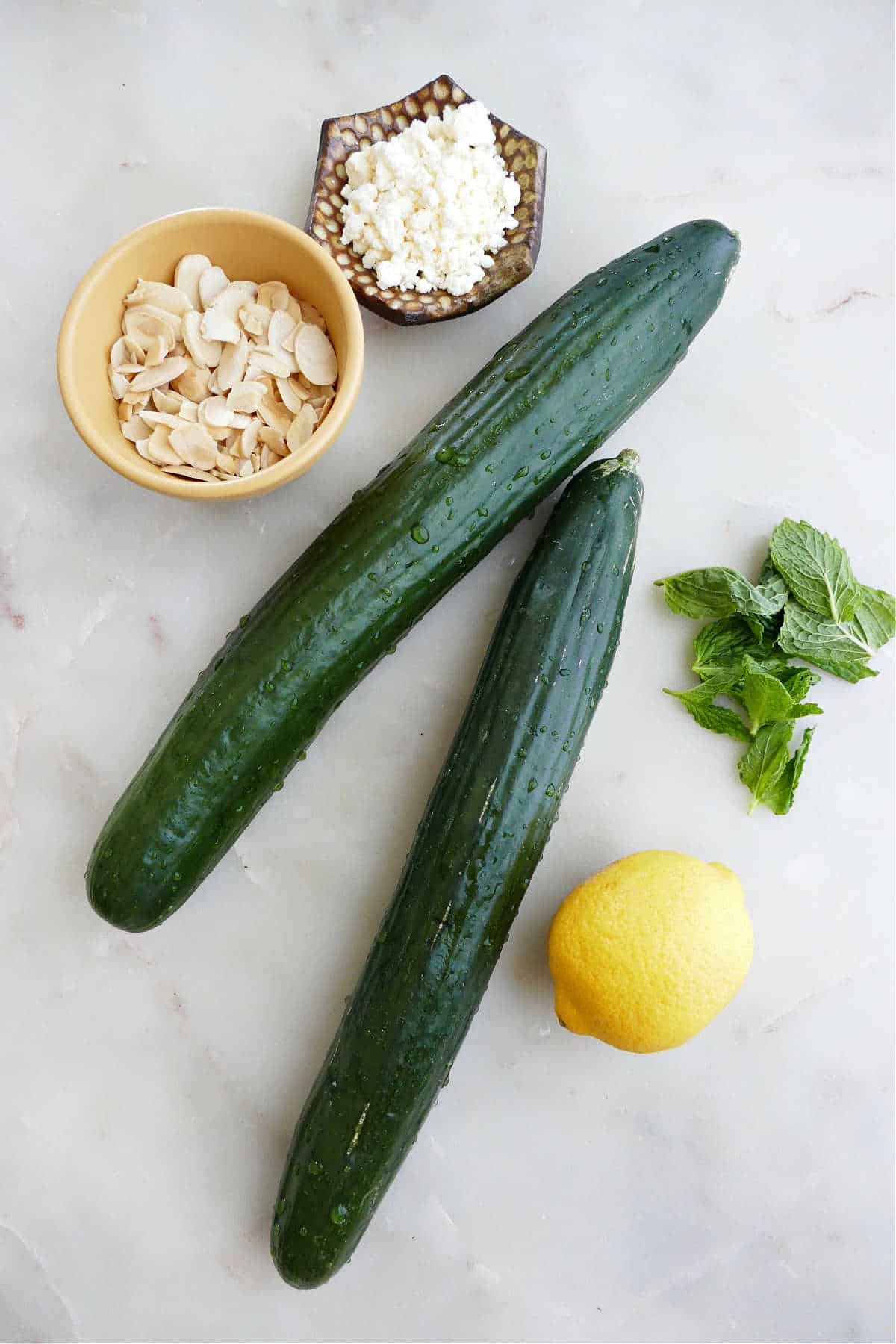 two cucumbers, a lemon, mint leaves, sliced almonds, and feta cheese on a counter