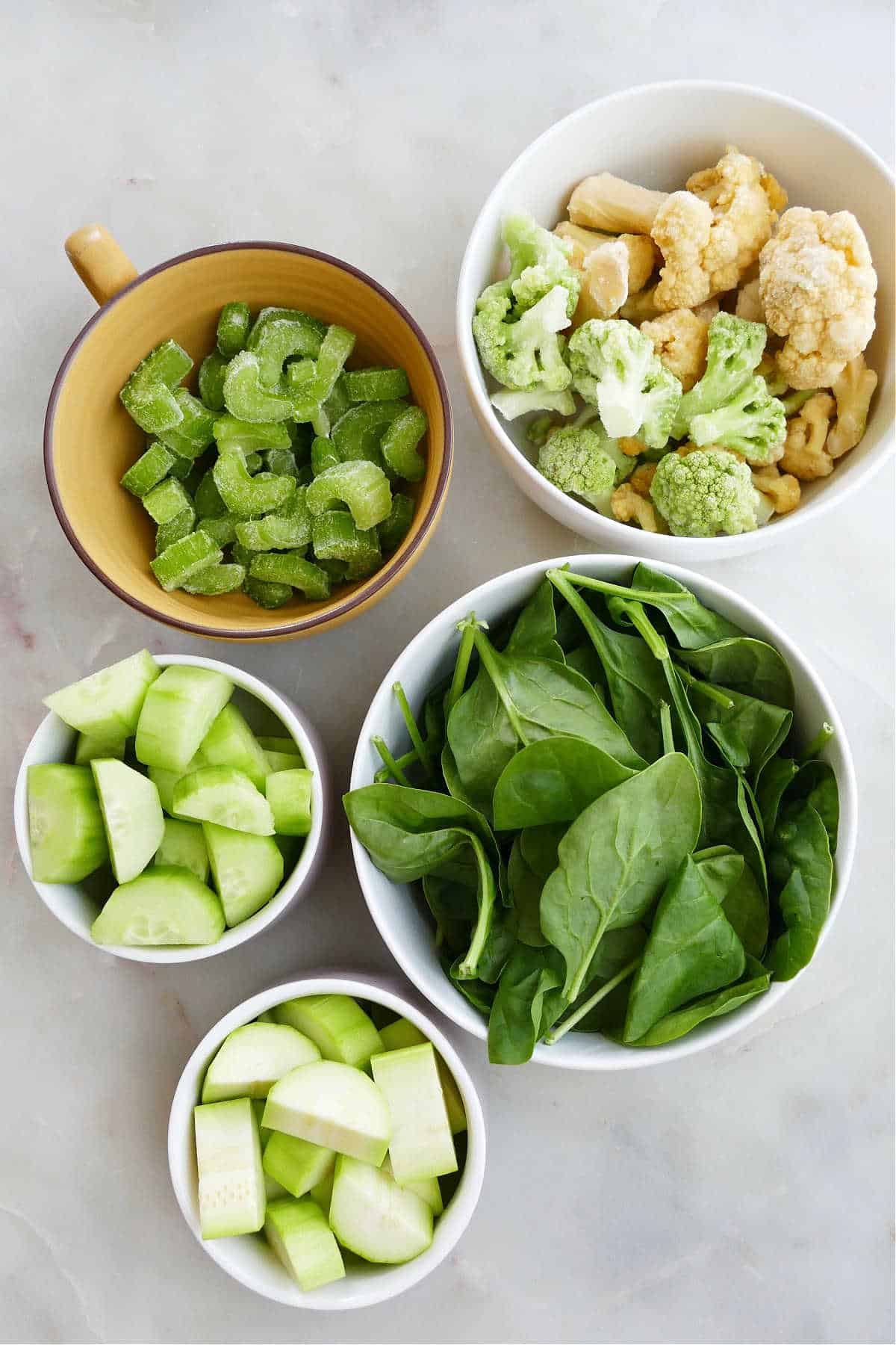 celery, cauliflower, spinach, cucumber, and zucchini in bowls spread out on a counter