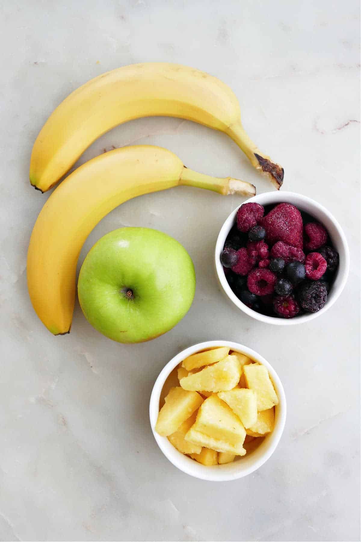 bananas, green apple, berries, and pineapple in bowls spread out on a counter