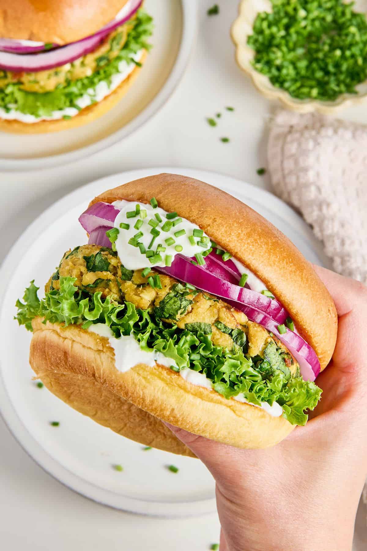 Woman holding a salmon burger with toppings in a bun over a plate.