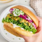 Woman holding a salmon burger with toppings in a bun over a plate.
