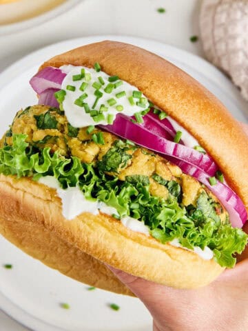Woman holding a salmon burger with toppings in a bun over a plate.