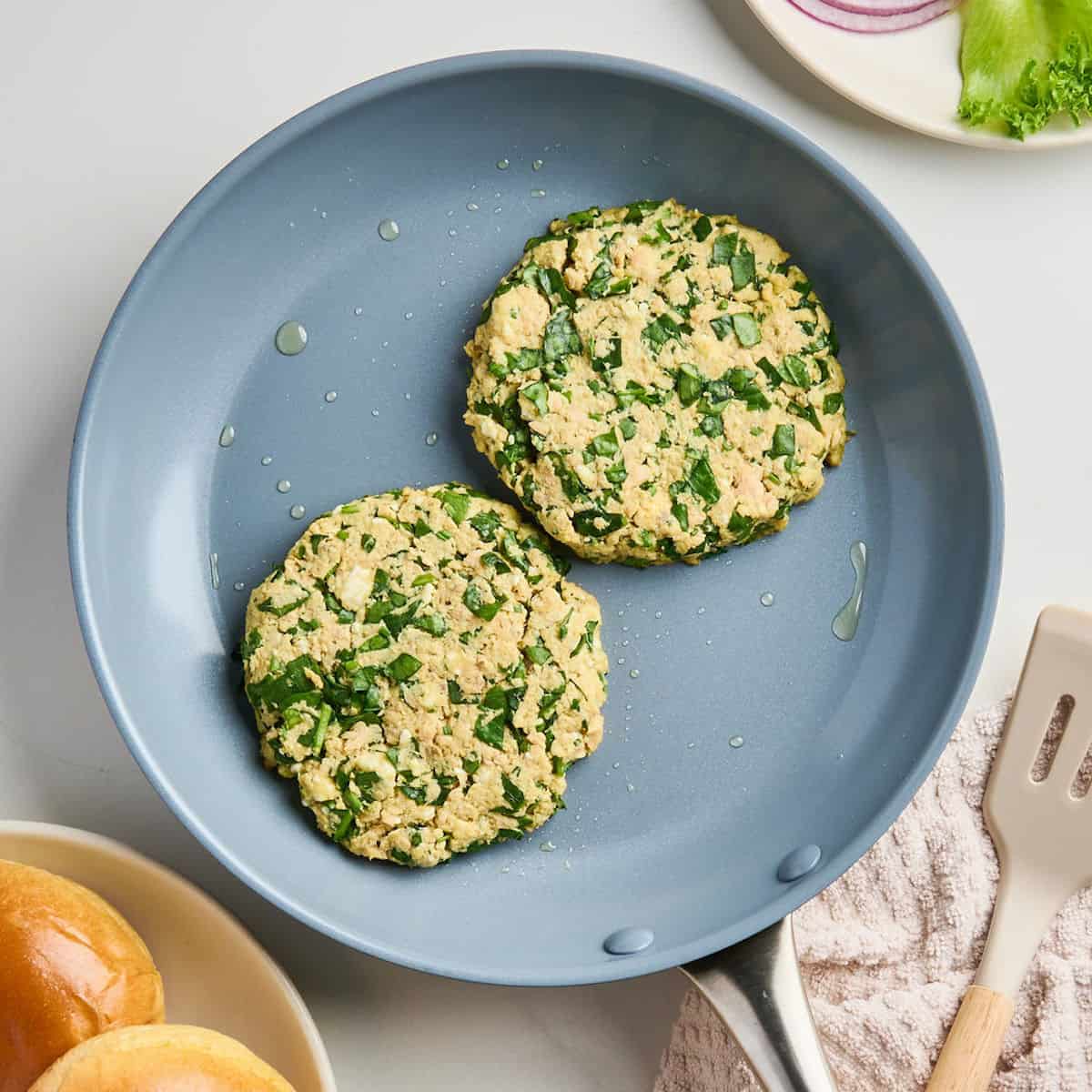Two salmon burgers cooking in a nonstick skillet in some oil next to ingredients.