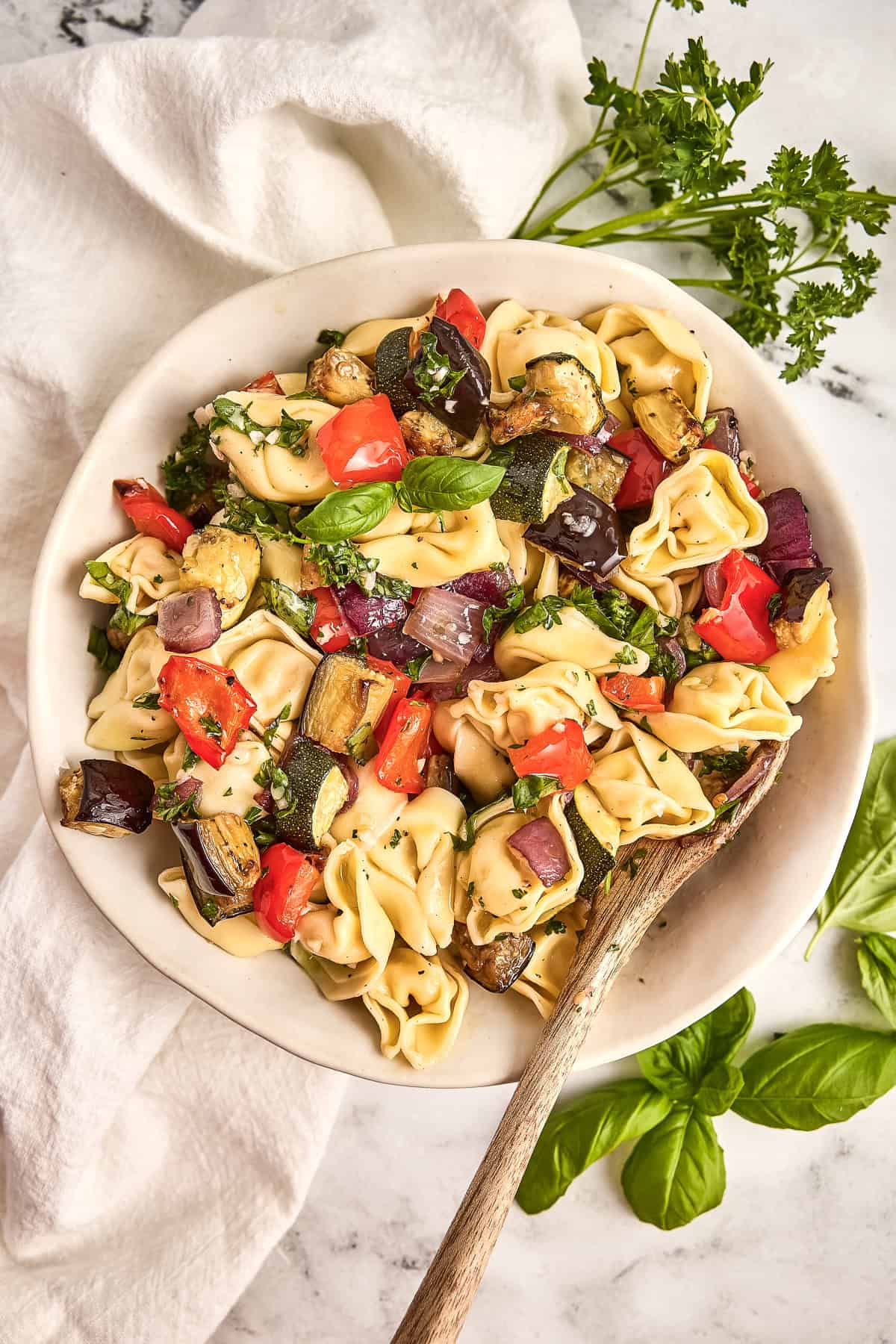Tortellini salad with roasted summer vegetables in a bowl with a spoon next to herbs.