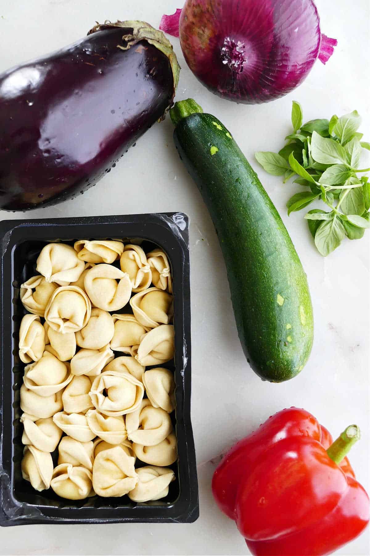 eggplant, red onion, zucchini, basil, red pepper, and tortellini spread out on a counter