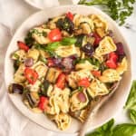Tortellini salad with roasted summer vegetables in a bowl with a spoon next to herbs.
