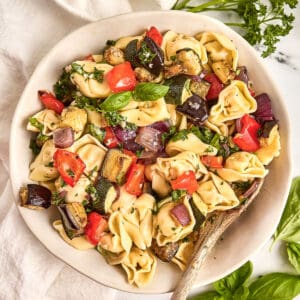 Tortellini salad with roasted summer vegetables in a bowl with a spoon next to herbs.