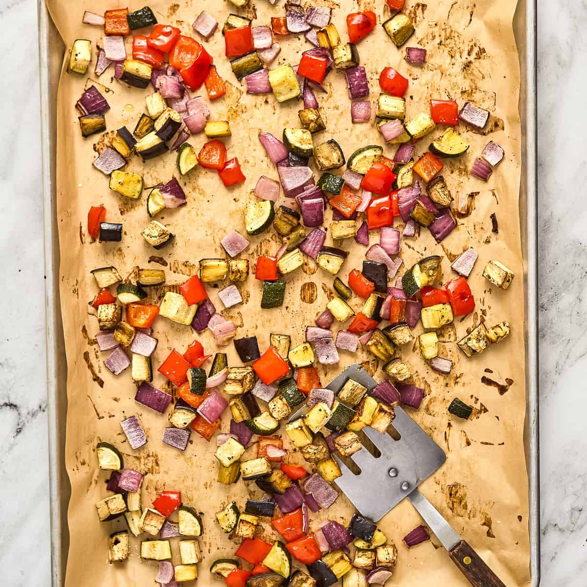 Roasted summer vegetables being scooped with a spatula on a baking sheet lined with parchment paper.