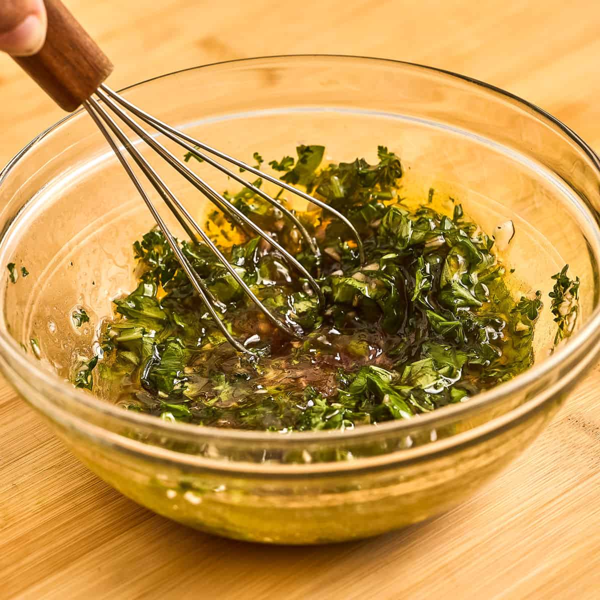 Woman whisking olive oil, vinegar, garlic, parsley, basil, and salt in a bowl.
