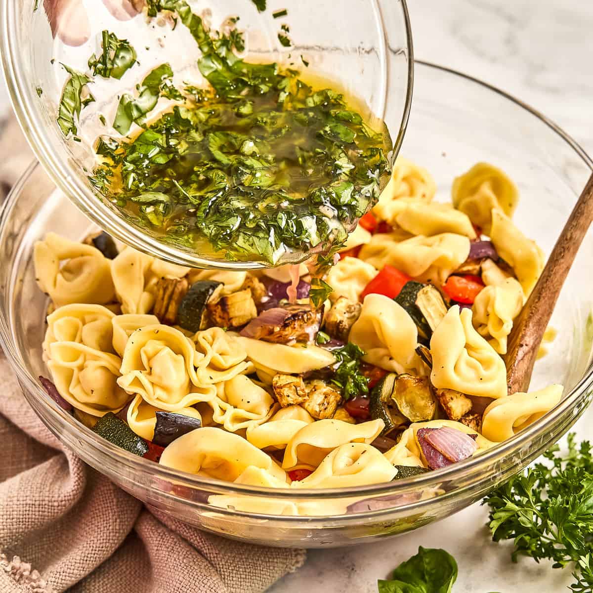 Woman pouring dressing over tortellini and roasted vegetables in a bowl with a spoon.