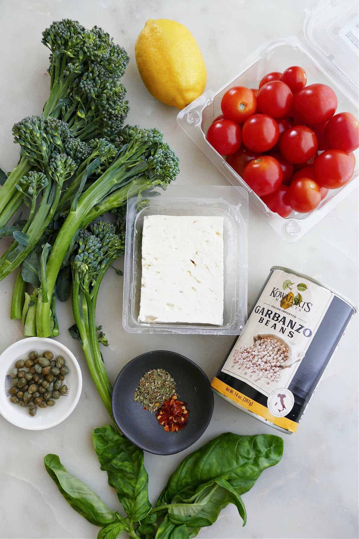 broccolini, lemon, tomatoes, feta, chickpeas, seasonings, capers, and basil on a counter