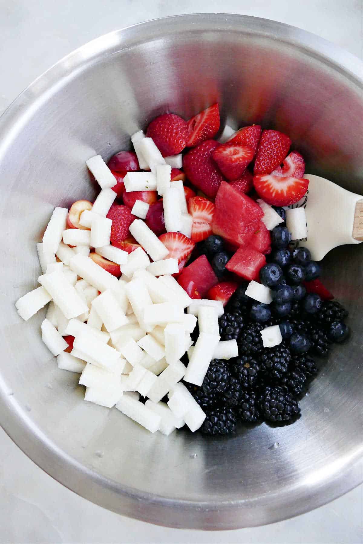 chopped watermelon, berries, and jicama in a mixing bowl with a rubber spatula