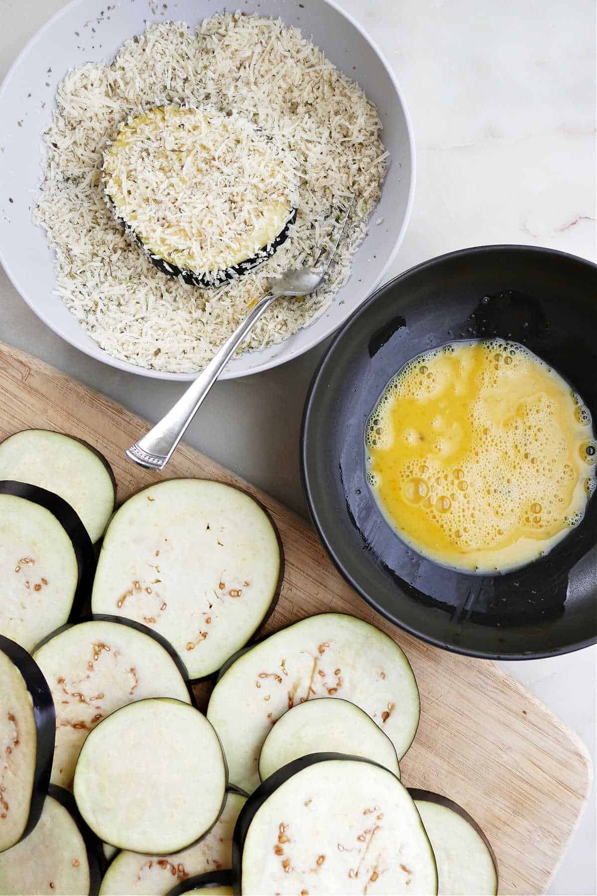 eggplant slices next to bowls with beaten eggs and breadcrumb coating