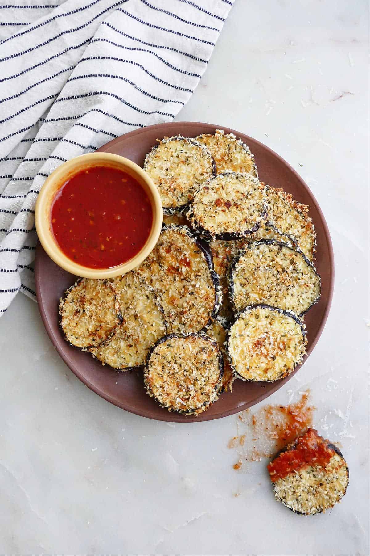 crispy baked eggplant slices on a serving dish with marinara sauce next to a striped napkin