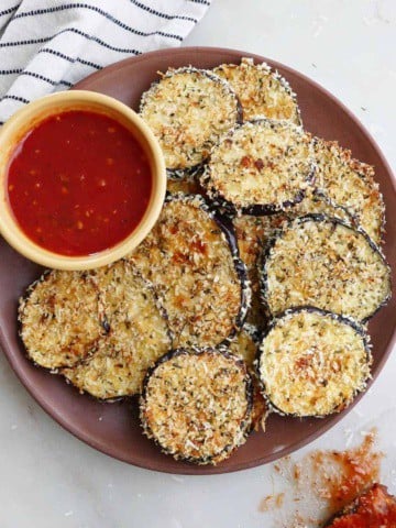 crispy baked eggplant slices on a serving dish with marinara sauce next to a striped napkin