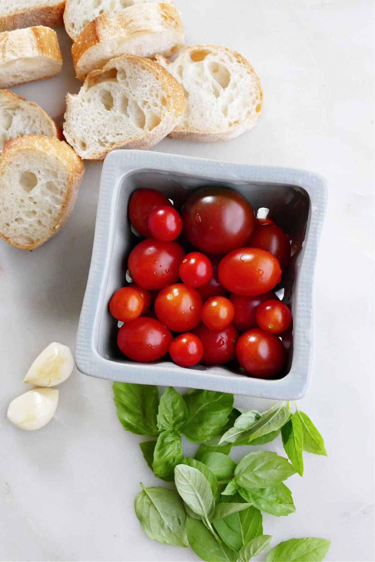 baguette slices, cherry tomatoes, garlic, and basil next to each other on a counter