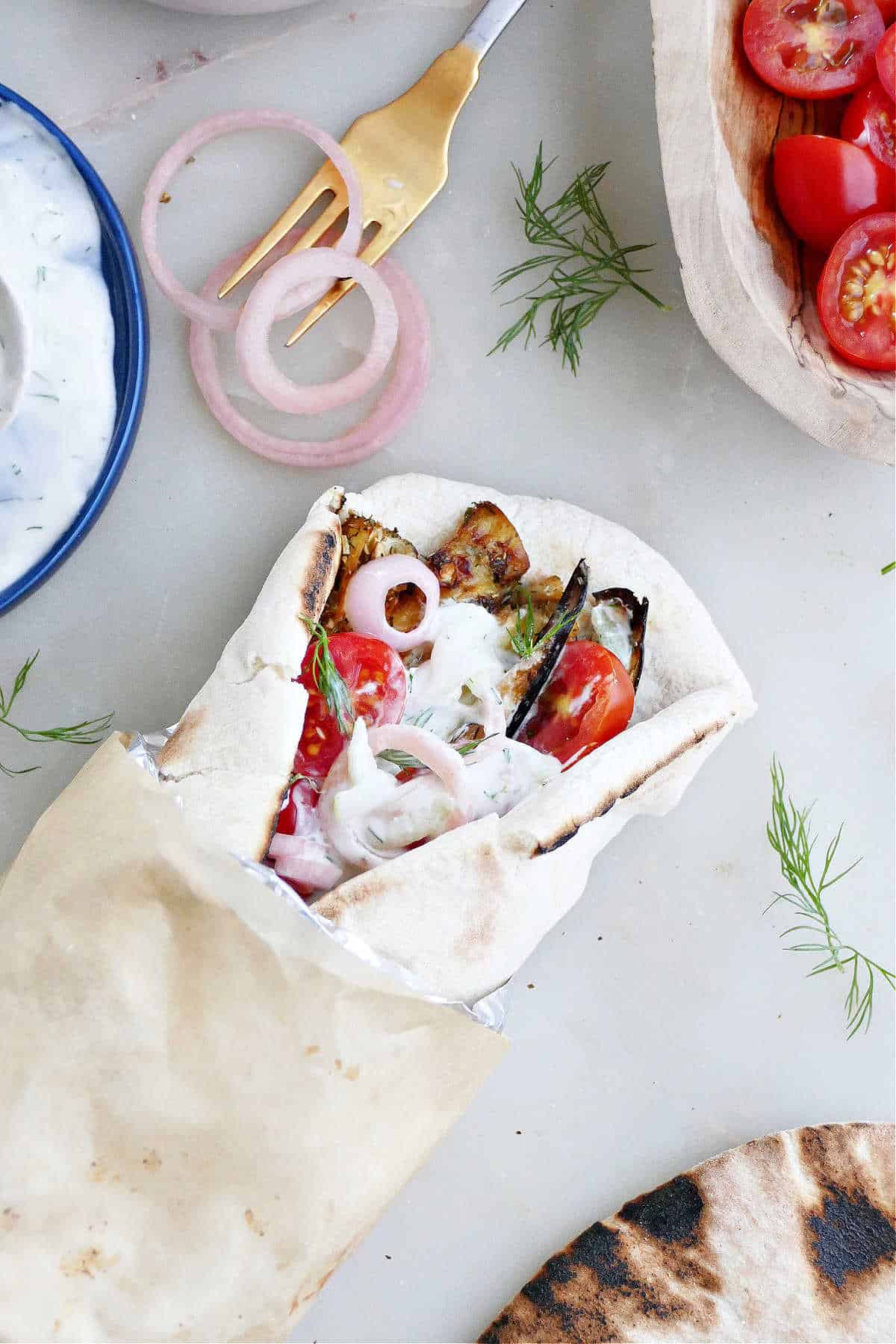 eggplant gyro sandwich on pita wrapped up in foil and parchment paper next to toppings