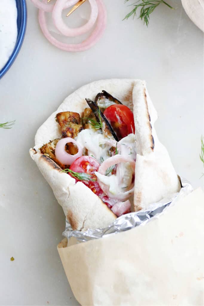 eggplant gyro sandwich on pita wrapped up in foil and parchment paper next to toppings