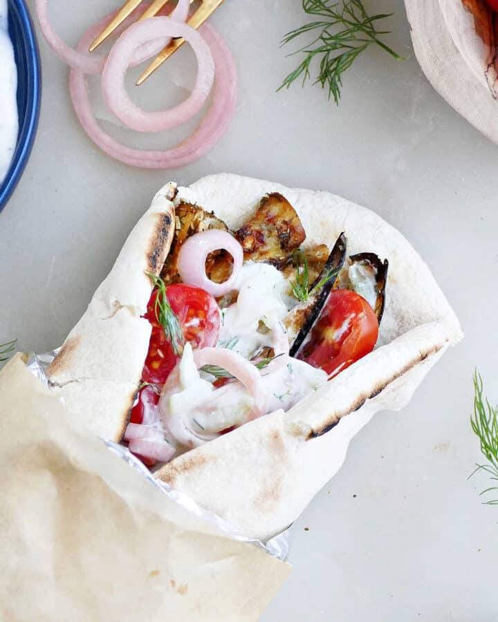 eggplant gyro sandwich on pita wrapped up in foil and parchment paper next to toppings