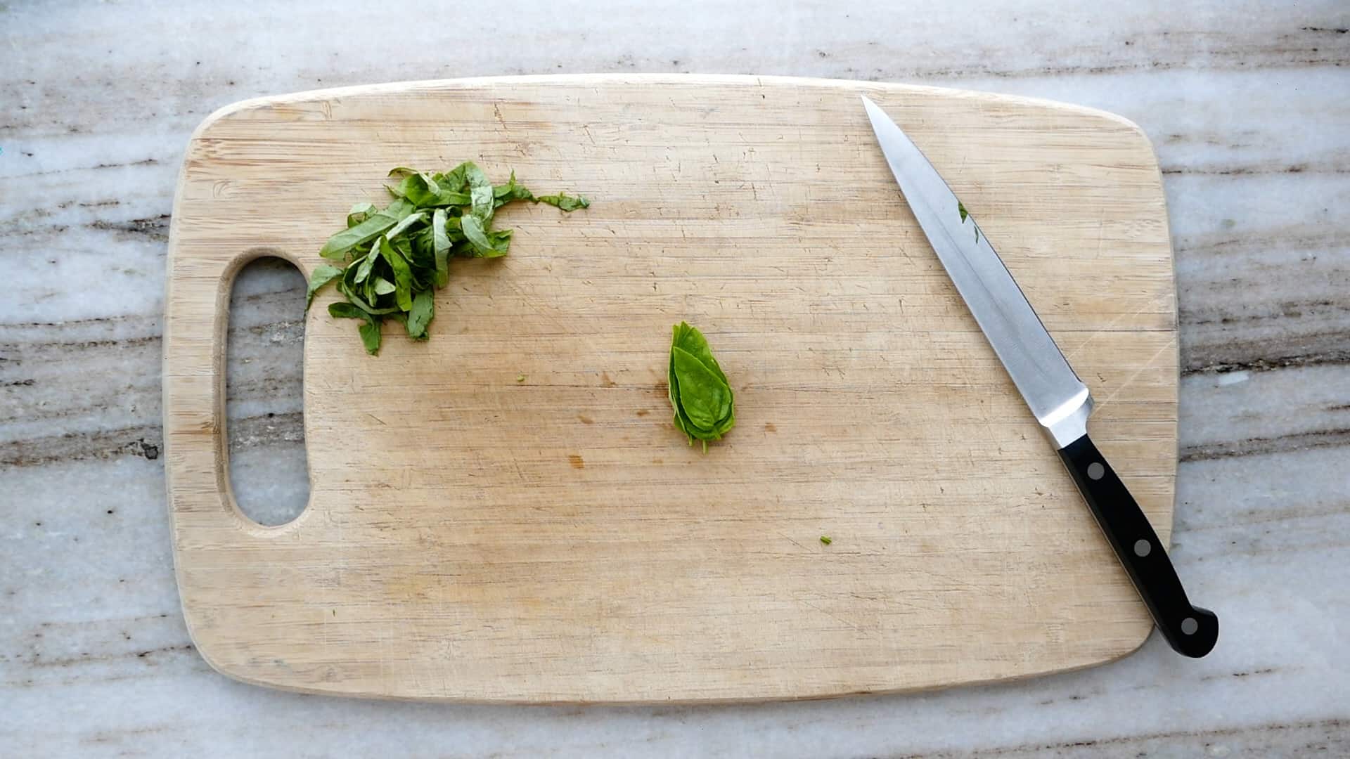 basil leaves in a stack in the center of a cutting board next to a knife