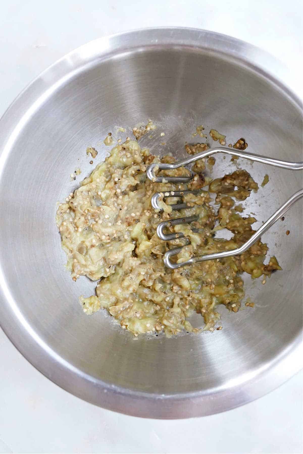 cooked eggplant being mashed in a mixing bowl with a potato masher