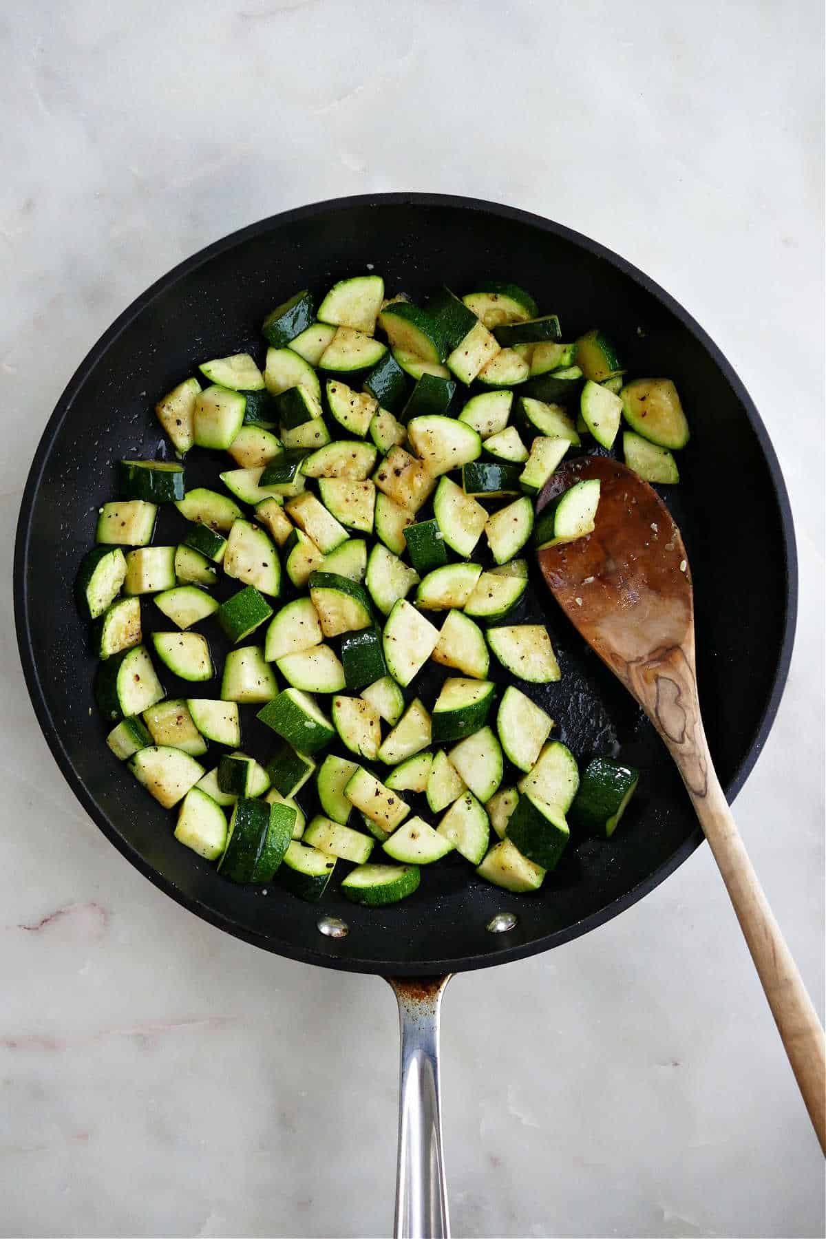diced zucchini cooking in a skillet with a wooden spoon in the pan