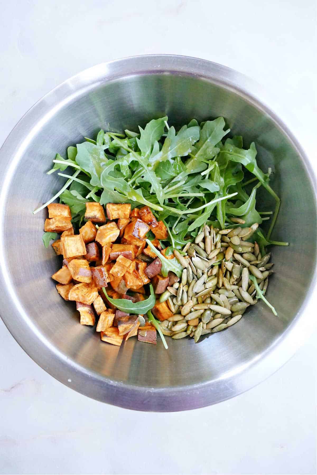 roasted sweet potato, arugula, and pumpkin seeds in a mixing bowl on a counter