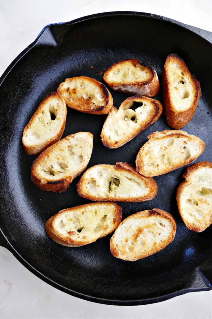 baguette slices brushed with oil and being toasted in a cast-iron skillet