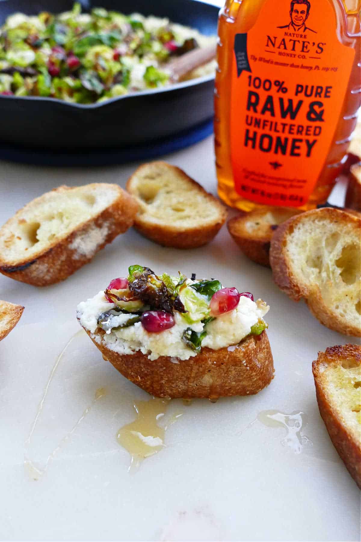 toasted baguette topped with ricotta dip, Brussels sprouts, and pomegranate arils in front of a honey bottle