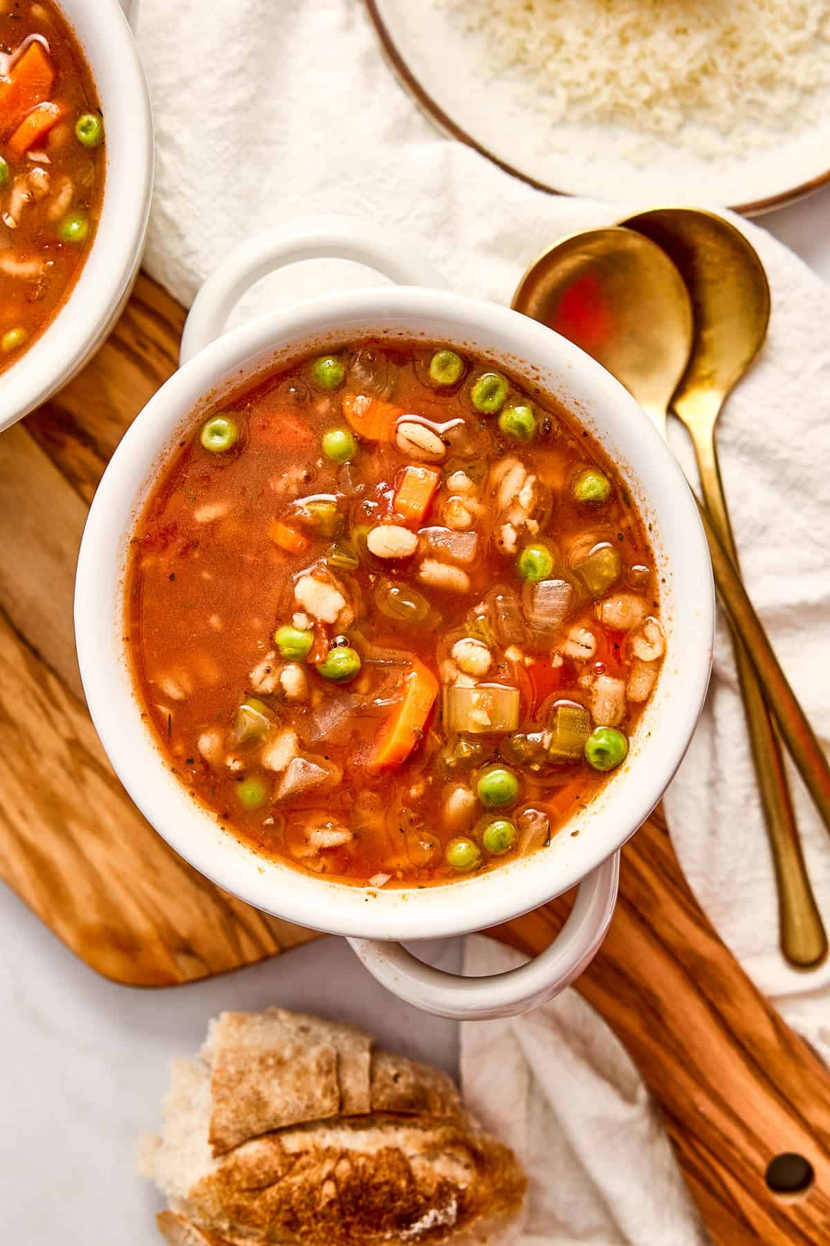 Bowl of vegetable barley soup next to bread, spoons, a napkin, and cheese.
