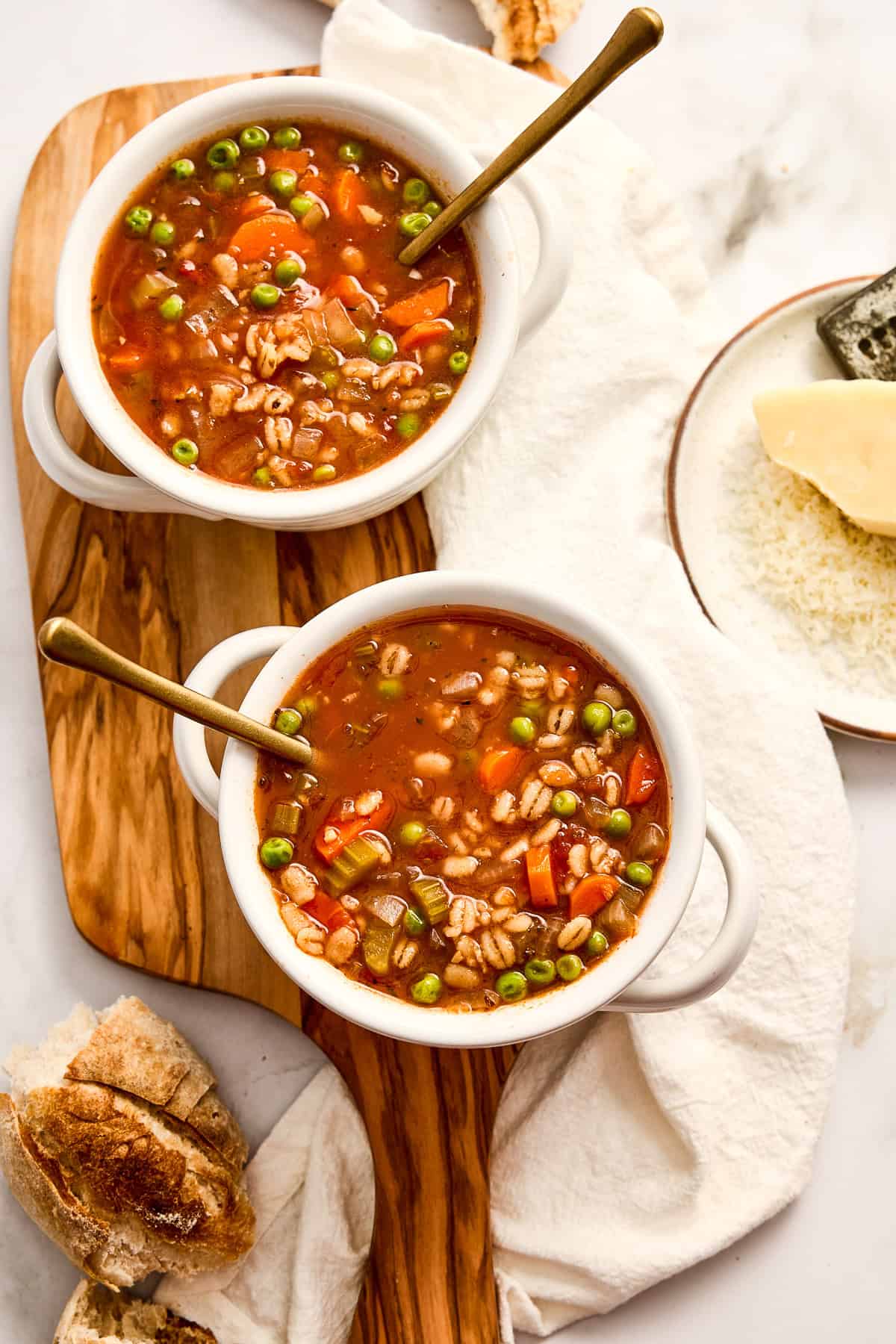 Two bowls of barley soup with vegetables on a board next to napkin, cheese, and bread.