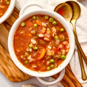 Bowl of vegetable barley soup next to bread, spoons, a napkin, and cheese.