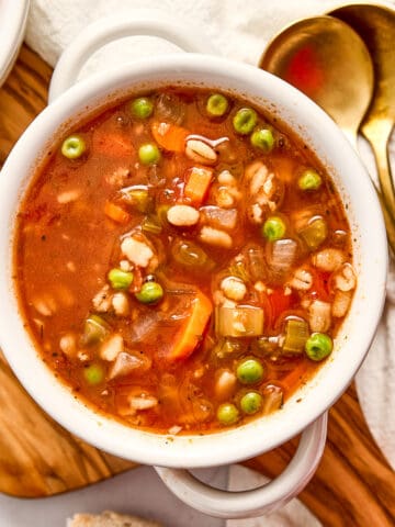 Bowl of vegetable barley soup next to bread, spoons, a napkin, and cheese.