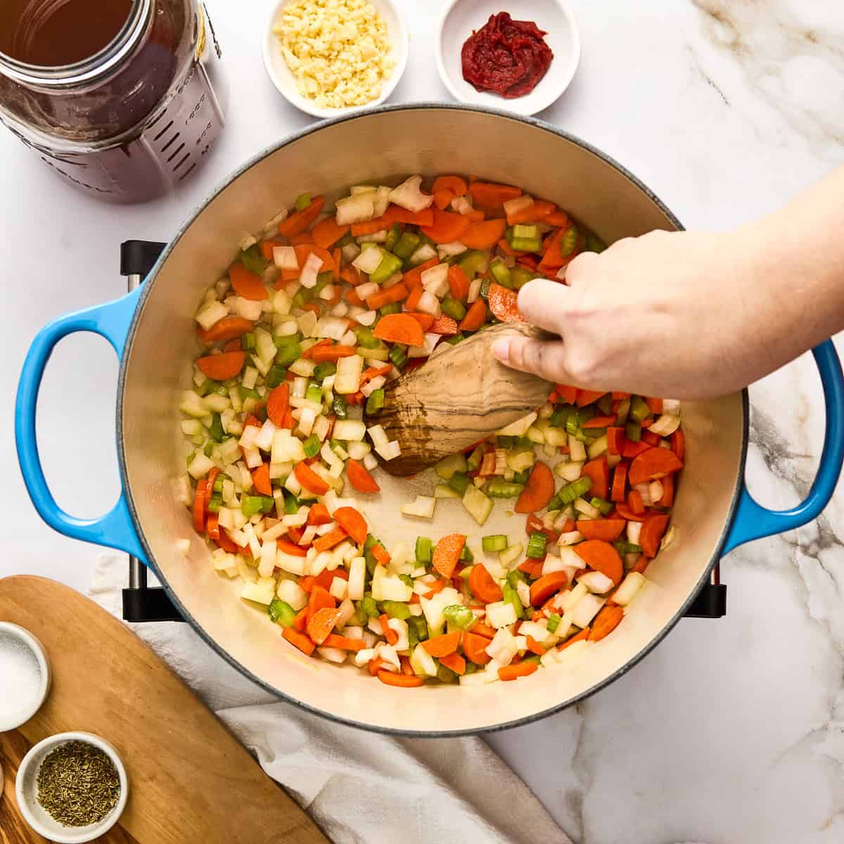 Woman stirring onion, celery, carrots, and garlic cooking olive oil in a soup pot.