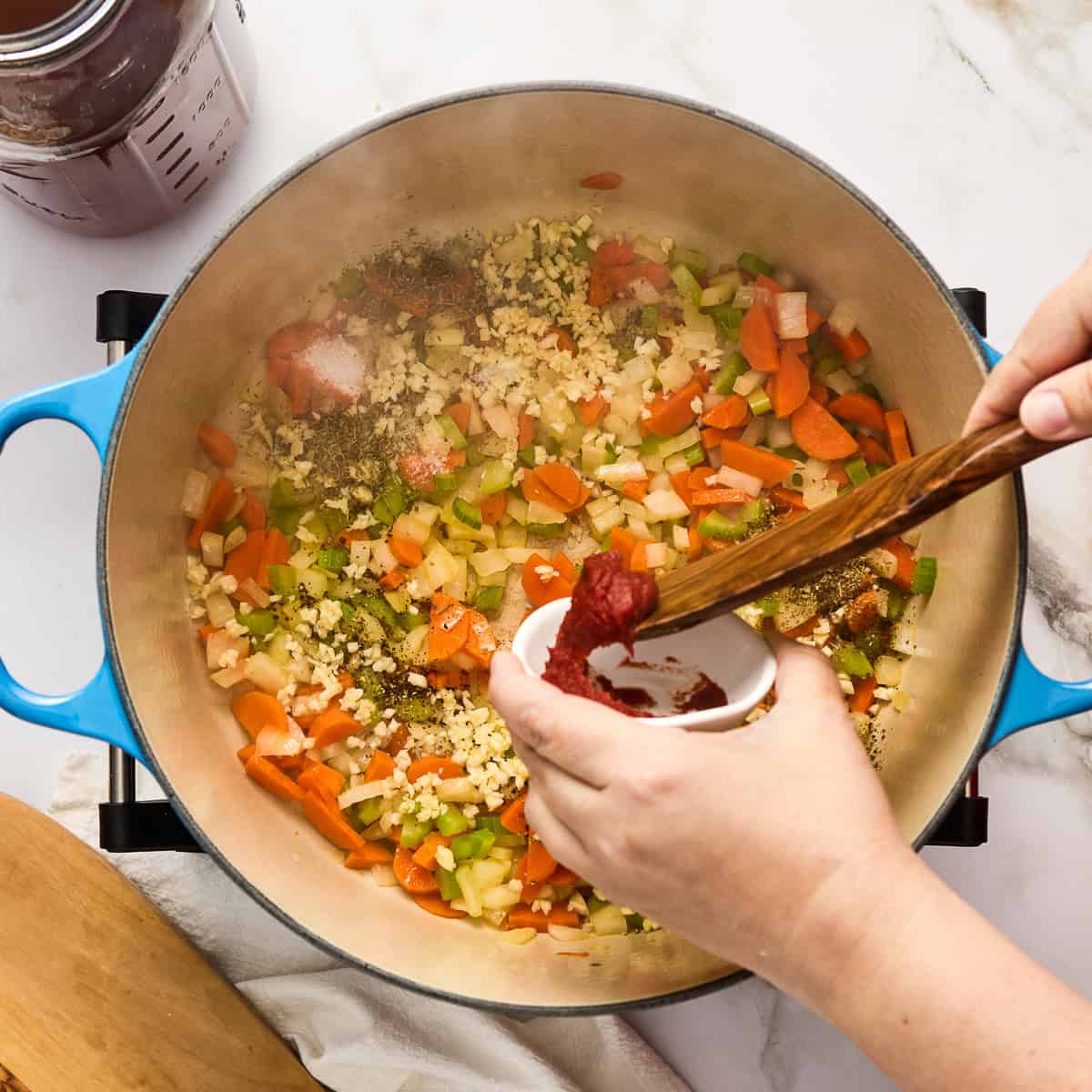 Woman adding tomato paste to a soup pot with vegetables cooking in oil.
