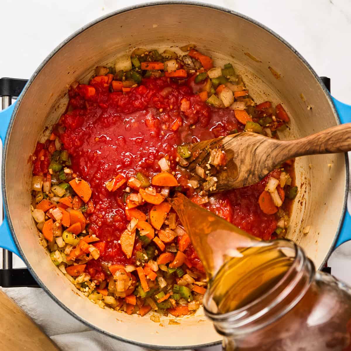 Woman pouring vegetable broth into a soup pot with mirepoix and tomatoes.