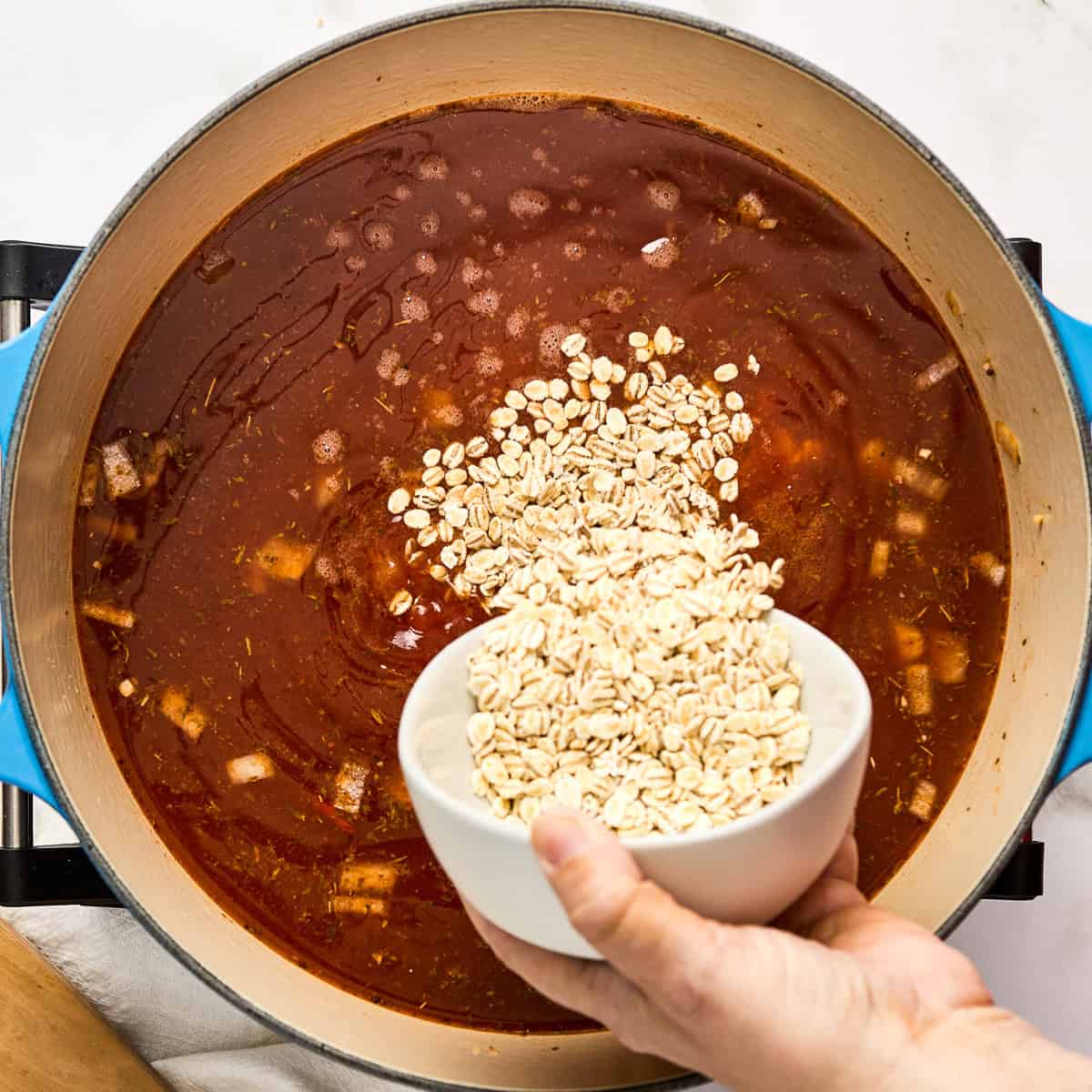Woman pouring barley into a soup pot with vegetables cooking in broth.