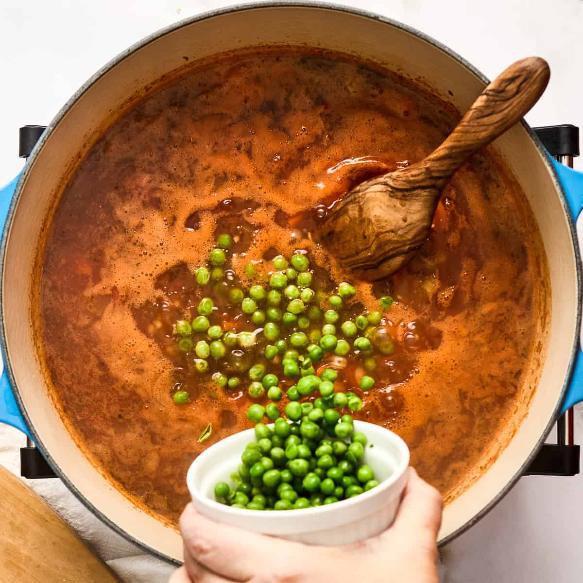 Woman adding peas to barley vegetable soup in a pot with a wooden spoon.