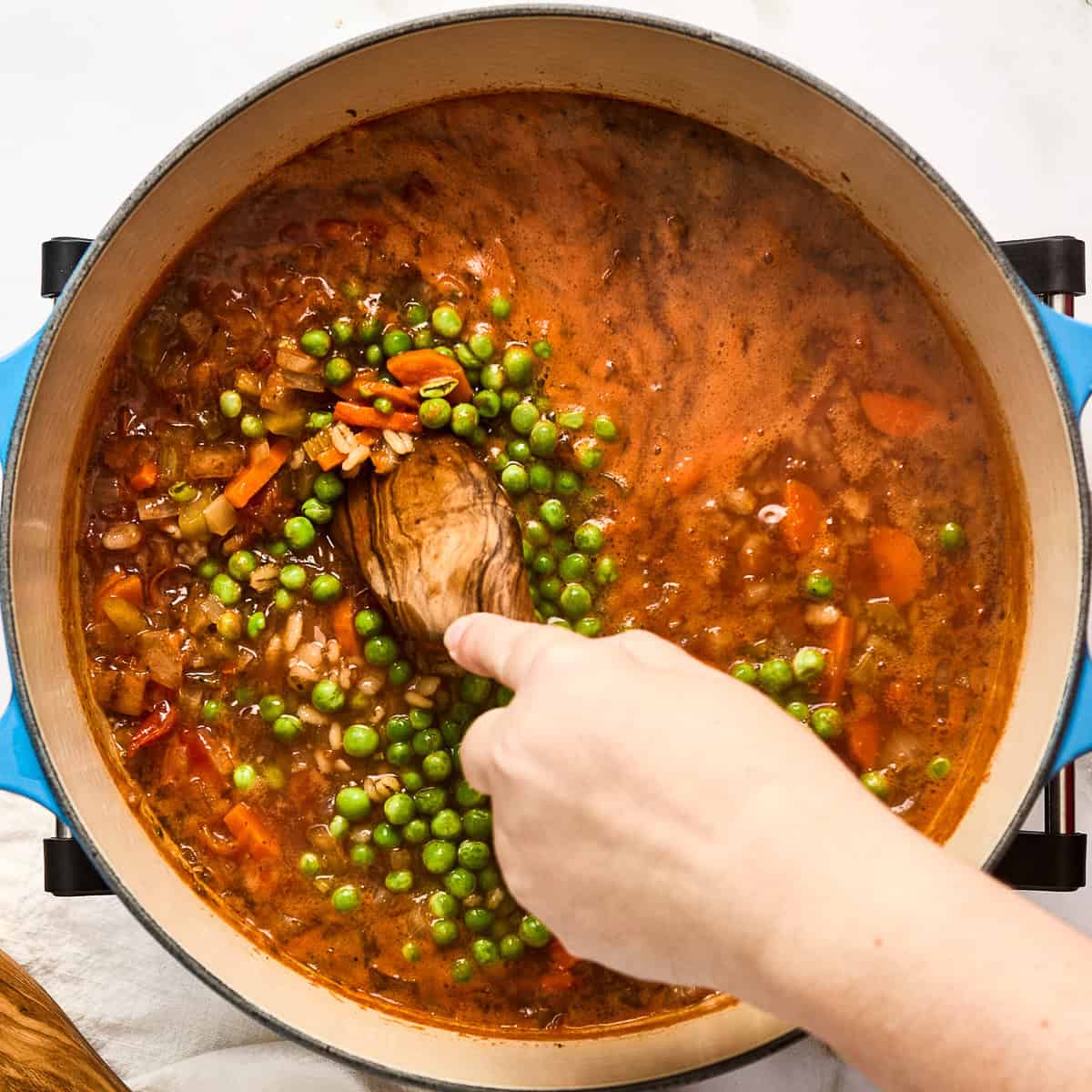 Woman stirring peas into barley vegetable soup cooking in a large pot.