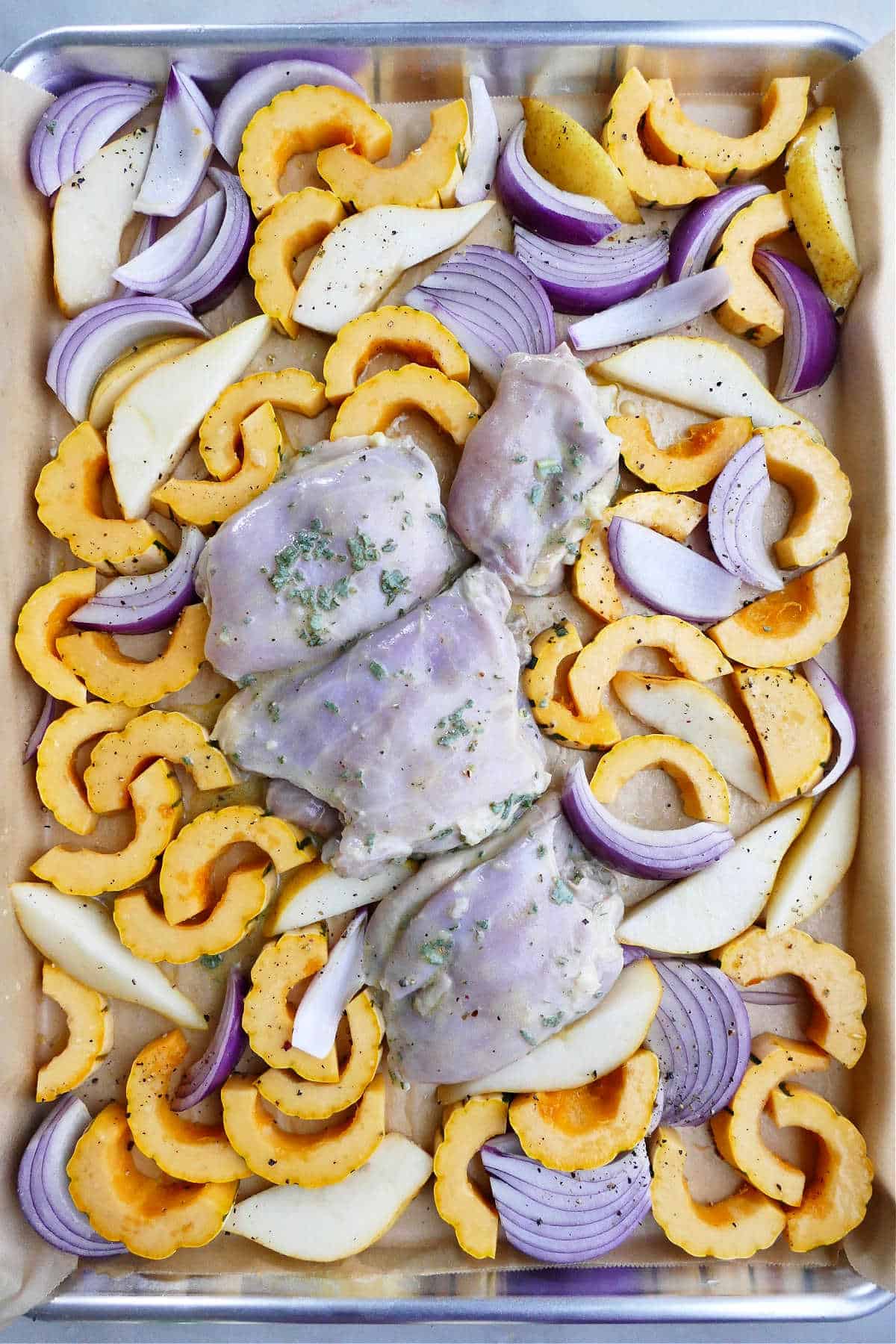 delicata squash, pears, onions, and chicken thighs spread out on a lined sheet pan