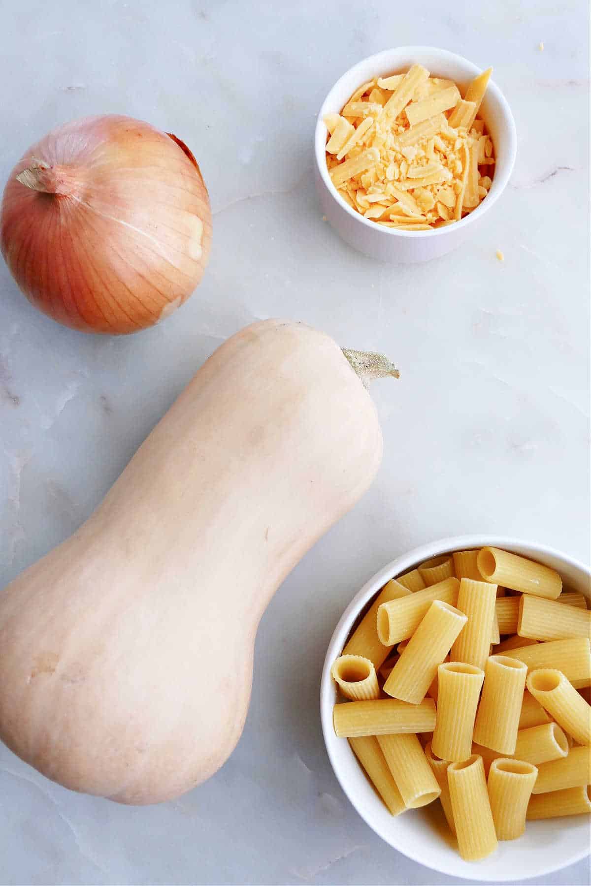 onion, butternut squash, shredded cheese, and rigatoni pasta on a counter next to each other