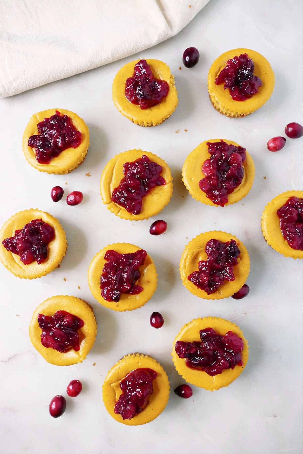 12 mini cranberry pumpkin cheesecakes on a counter next to napkin and whole cranberries