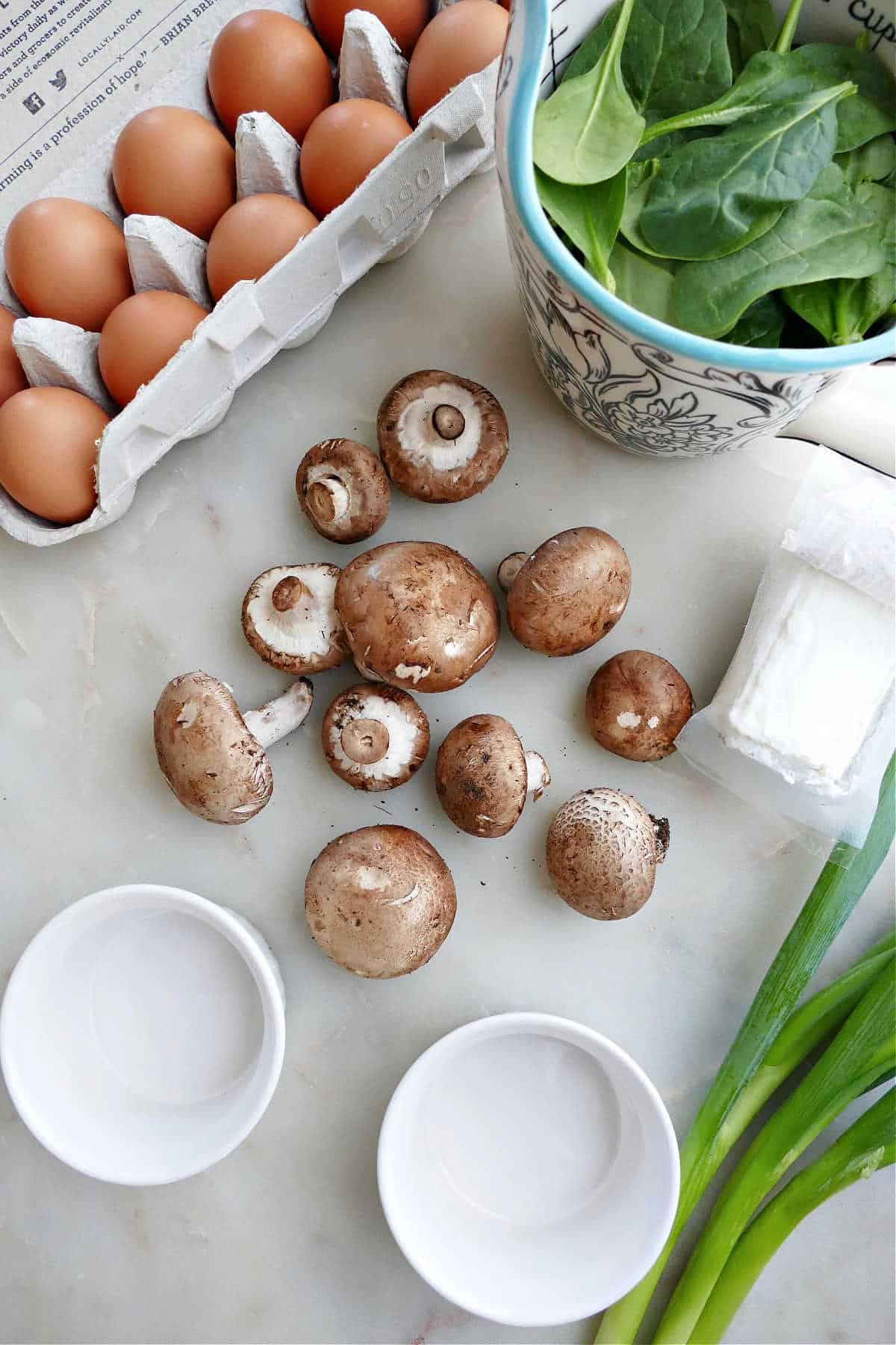 eggs, mushrooms, spinach, goat cheese, and scallions on a counter next to ramekins