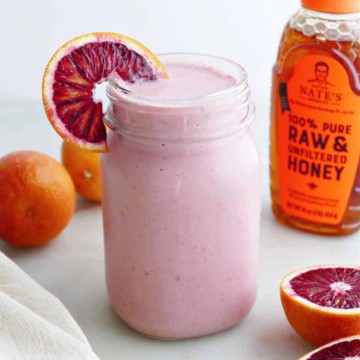 blood orange smoothie in a glass with an orange wedge in front of honey bottle