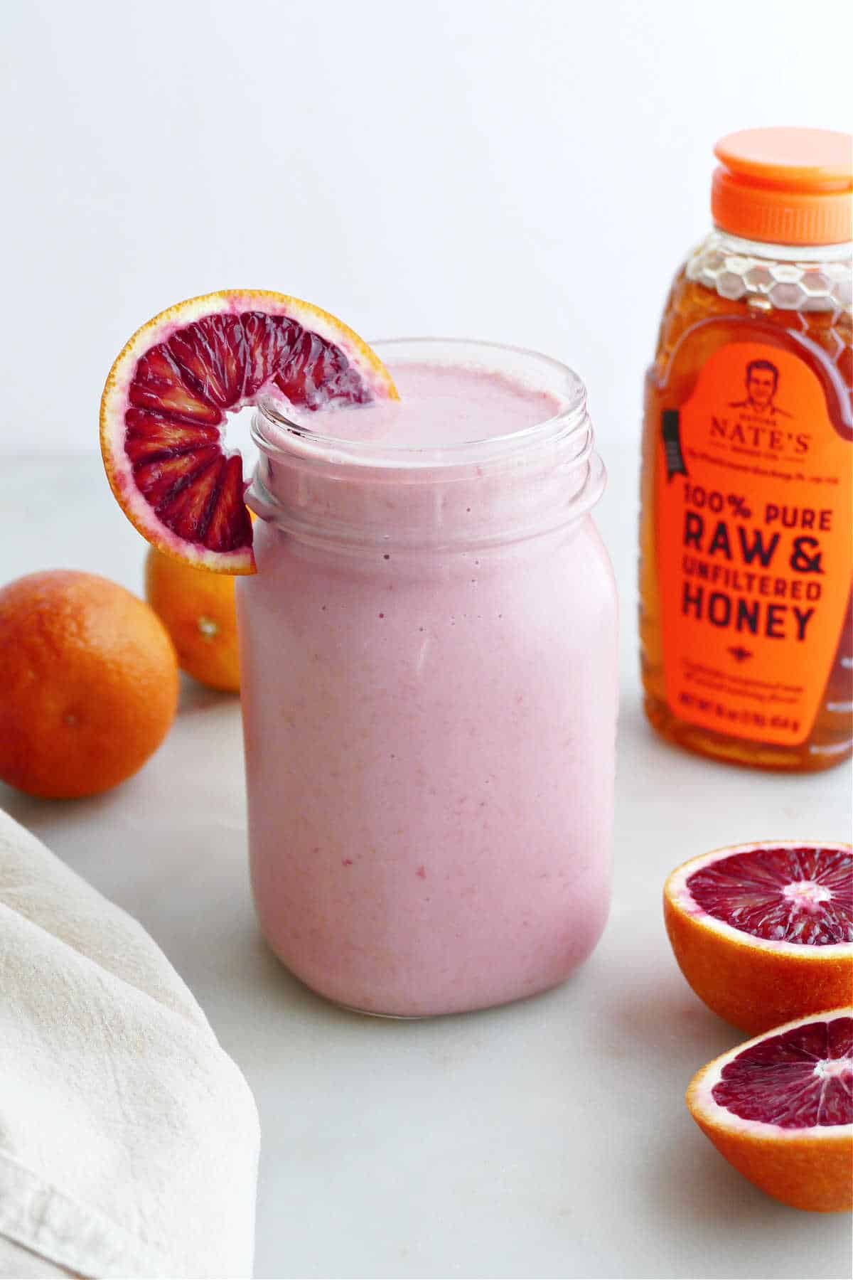 blood orange smoothie in a glass with an orange wedge in front of honey bottle