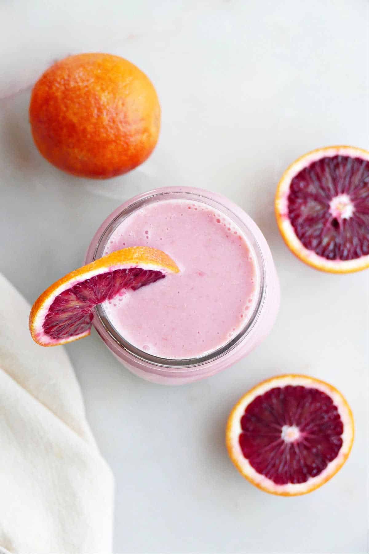 blood orange smoothie in a glass with a wedge on the rim surrounded by blood oranges