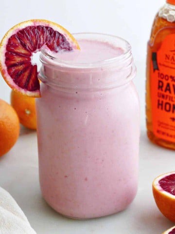 blood orange smoothie in a glass with an orange wedge in front of honey bottle